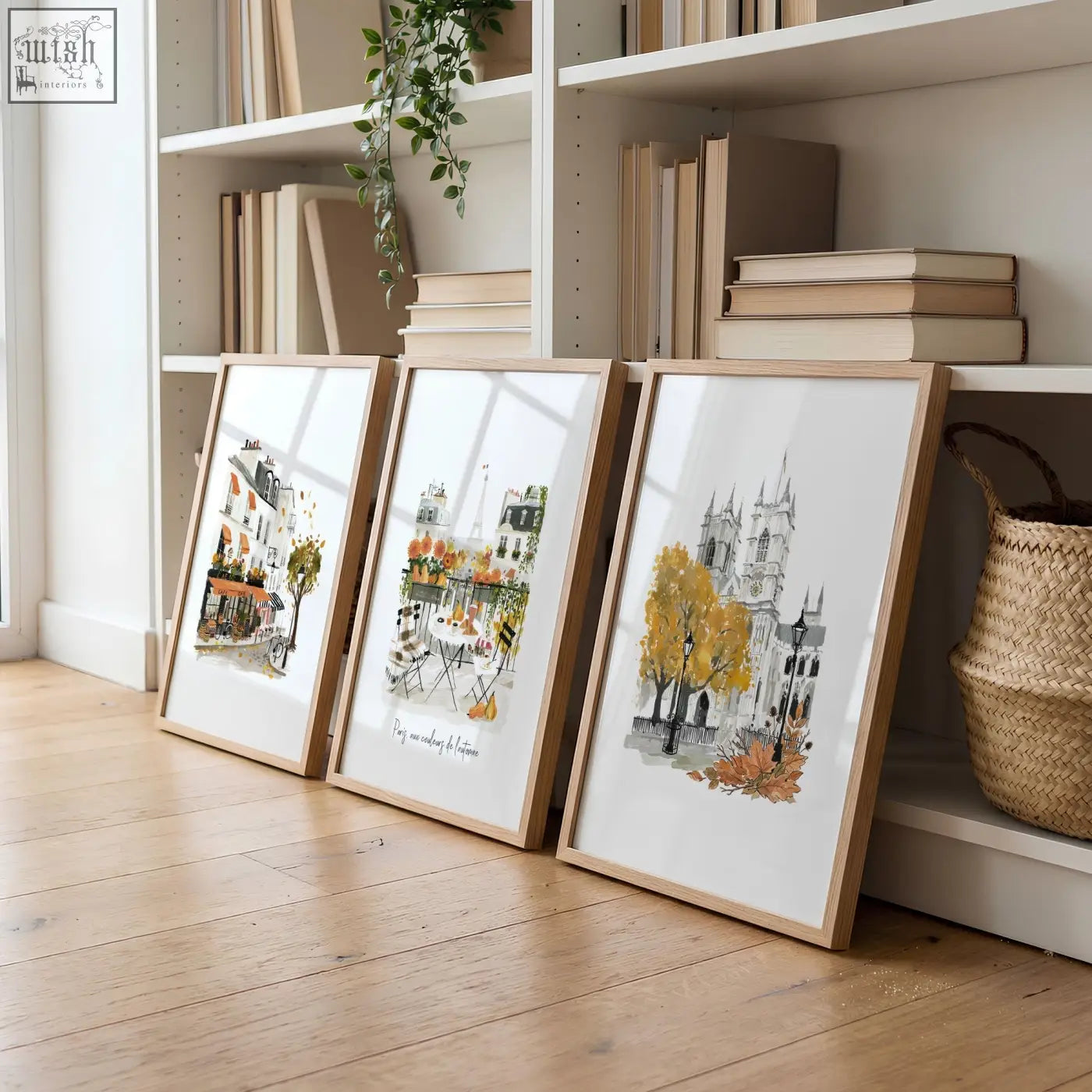 Three large artwork prints of autumn paris watercolour in light wood frames leaning against a white bookshelf in living room