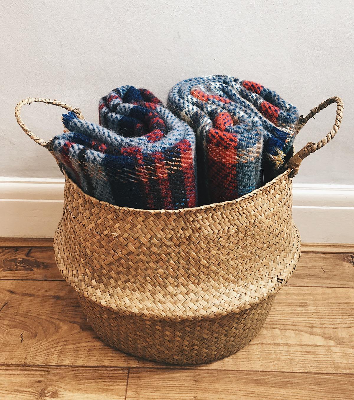 A woven basket holding rolled-up large checked random recycled wool blankets in blue red and black shades
