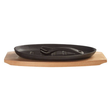 Black oval cast iron sizzler dish with built-in lifting handle resting on wooden trivet