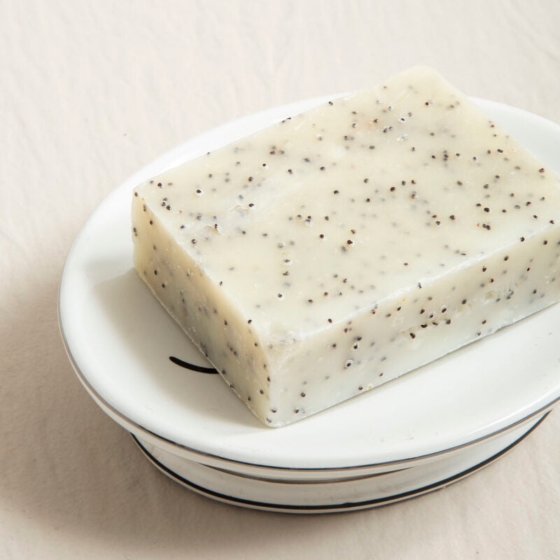 A rectangular off-white bar soap with black poppy seed specks on a le bain white ceramic soap dish