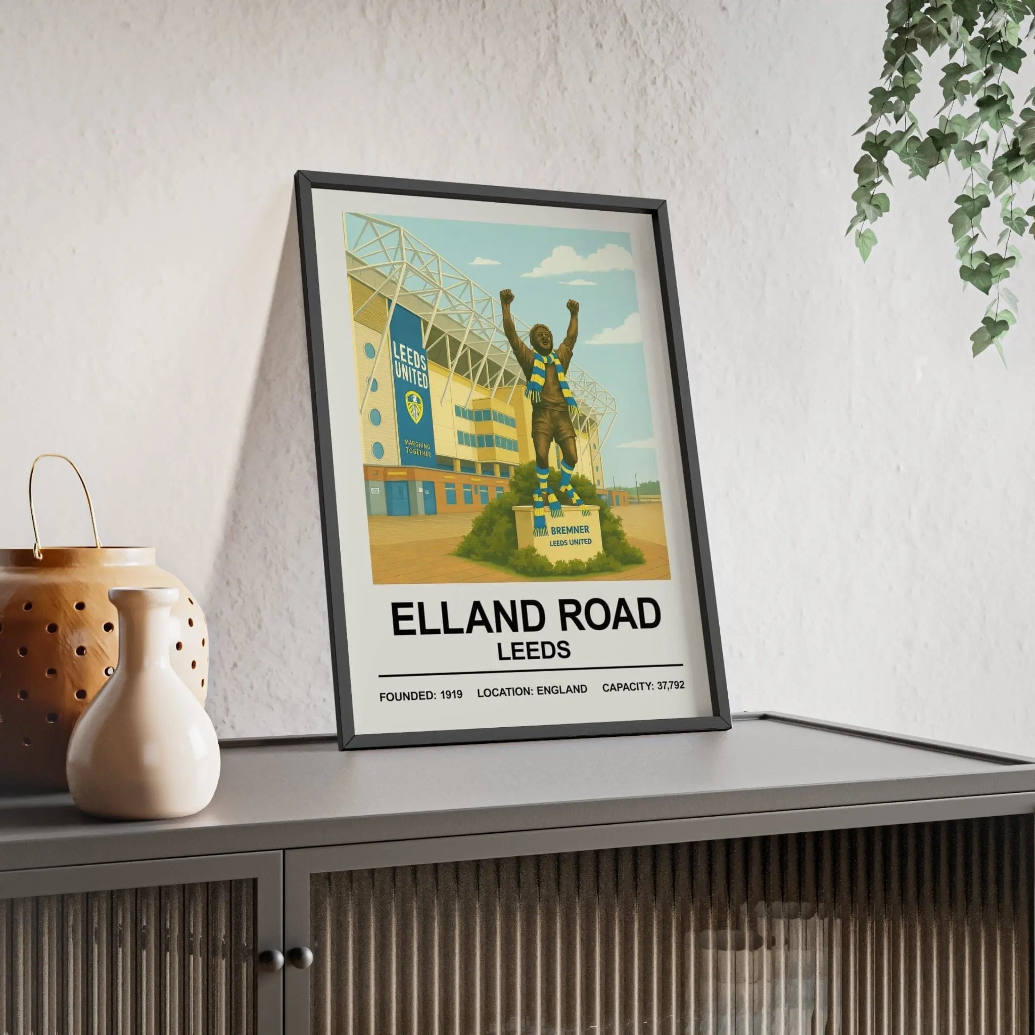 A framed vintage-style print of elland road leeds stadium with a bronze statue wearing a blue and white scarf in front