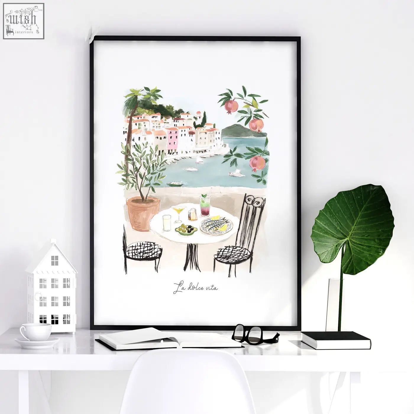 A framed watercolor living room artwork showing a mediterranean coastal café with pastel buildings and pomegranates