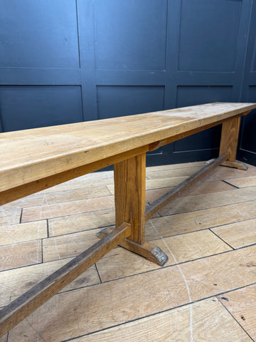 A rustic light oak wooden bench with a long rectangular top and sturdy t shaped legs for leys school seating