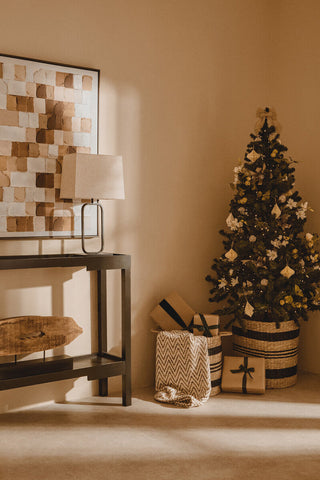 A dark green christmas tree with white and gold ornaments in a woven basket beside a lucas natural and black table lamp