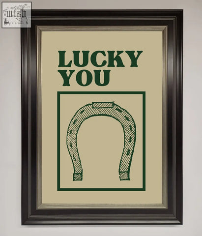 A framed wall art print with a green horseshoe and lucky you text on an off-white background