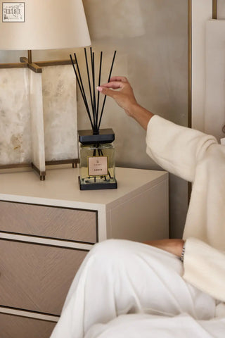 A sleek black luxury square reed diffuser with a clear glass container and gold-toned ambra scent label