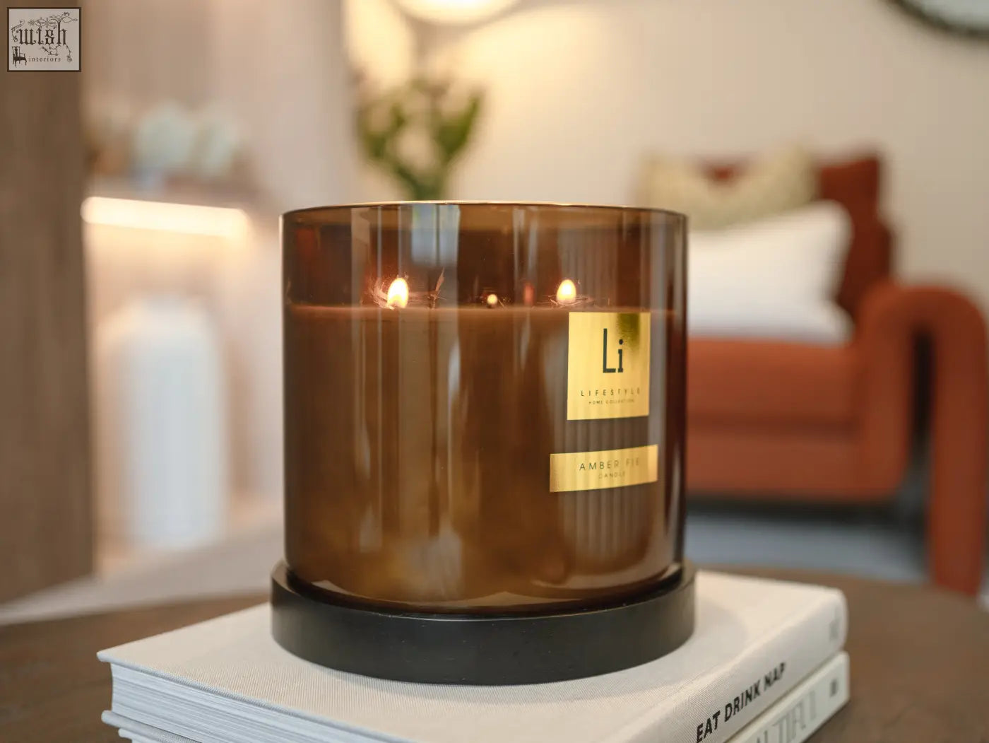 A tall amber fie luxury xxl candle with three lit wicks and gold label on black base over stacked books
