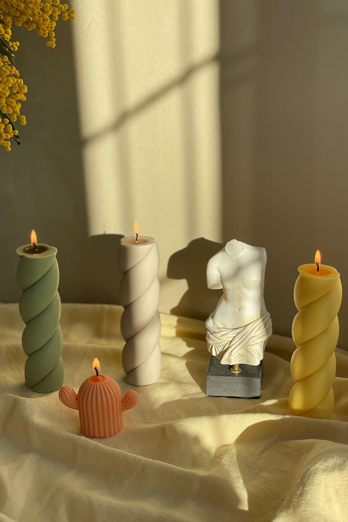 A white marshmallow shaped pillar candle with a twisted design and lit wick on a soft fabric surface