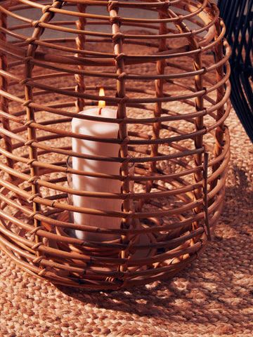 A tall natural rattan lantern in warm tones housing a white candle perfect for cozy lighting