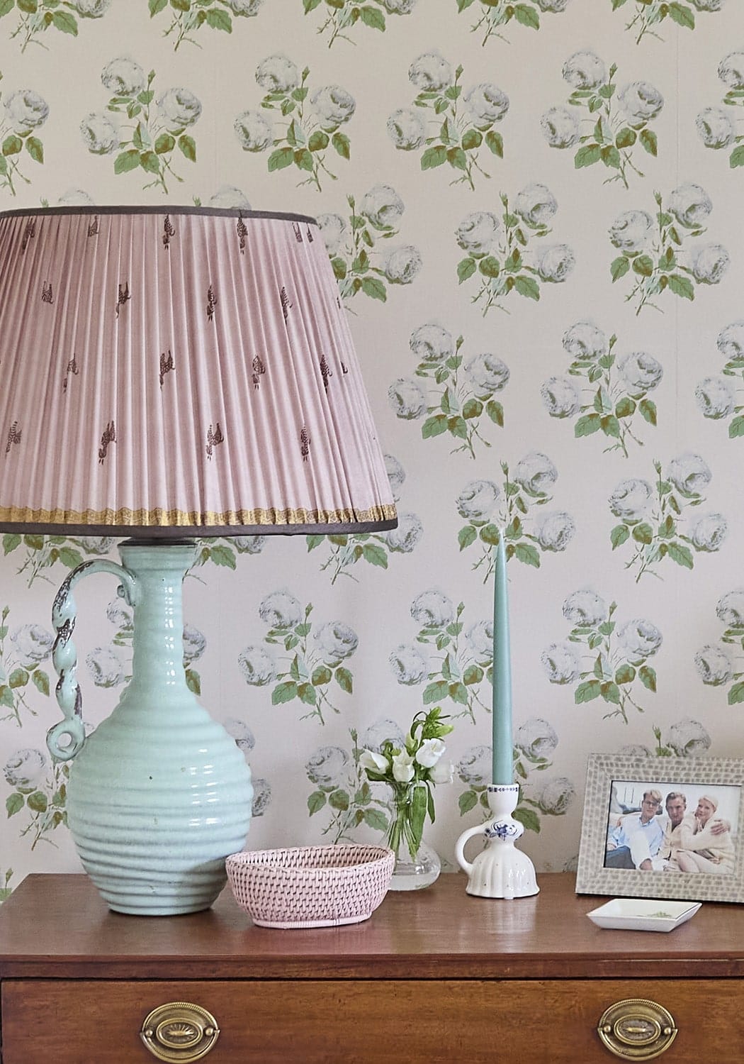 A delicate pink pleated lampshade with gold trim atop a mint green ceramic lamp with ornate handle next to a pink mayfair trinket hold all rattan basket