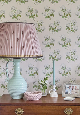 A delicate pink pleated lampshade with gold trim atop a mint green ceramic lamp with ornate handle next to a pink mayfair trinket hold all rattan basket