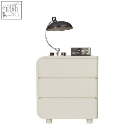 Cream-colored meg bedside table with three drawers and rounded edges topped by black lamp and digital clock clean with damp cloth