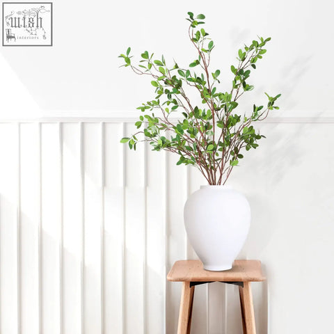 A sleek white ceramic vase holding green faux branches from melorca&guilla artificial plants set for home decor