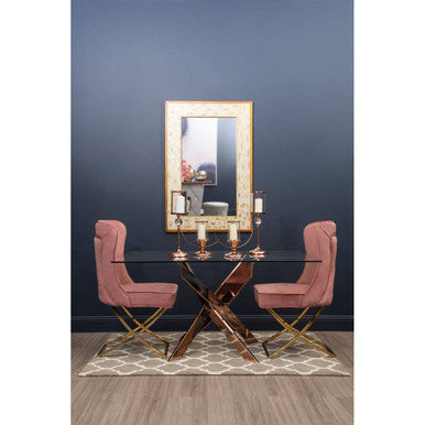 A modern dining area with a meril rectangular wall mirror above a glass top table and pink velvet chairs against dark blue wall