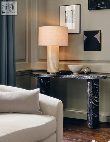 Cream-colored cylindrical table lamp with textured fabric shade and polished stone base on midnight luxe black console table with natural marble surface