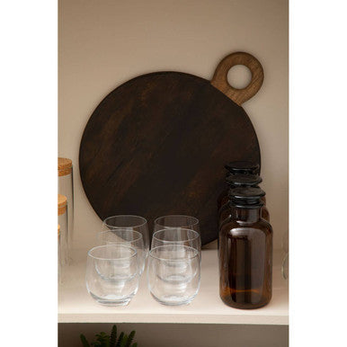 Dark brown wooden cutting board with circular handle next to mila set glass tumblers with a wide bowl design