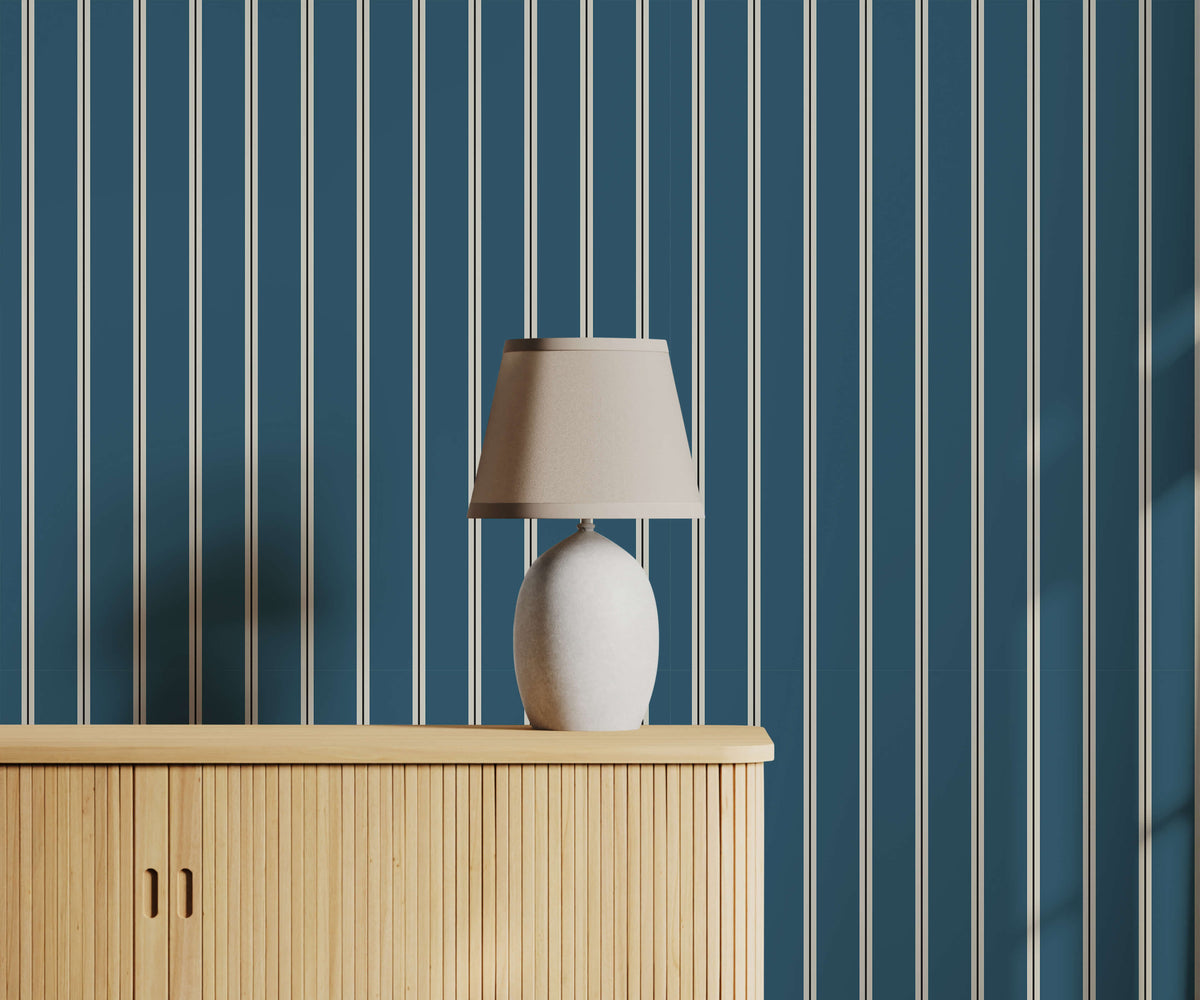 A sleek white ceramic miller table lamp with tapered beige fabric shade on light wood cabinet with pinstripe wallpaper