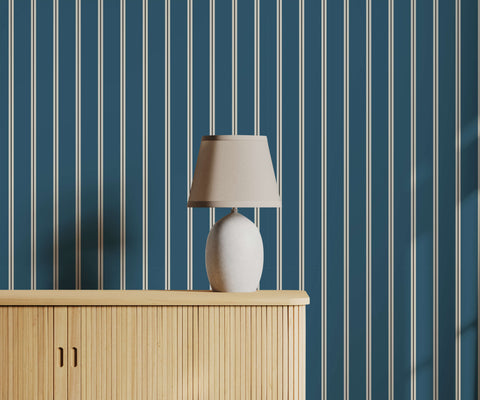 A sleek white ceramic miller table lamp with tapered beige fabric shade on light wood cabinet with pinstripe wallpaper