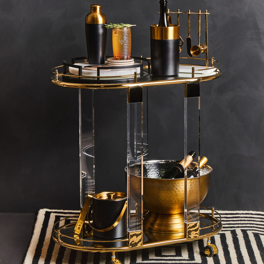 Gold and black two-tiered bar cart with clear acrylic legs and gold finish ice bucket on gold-rimmed trays