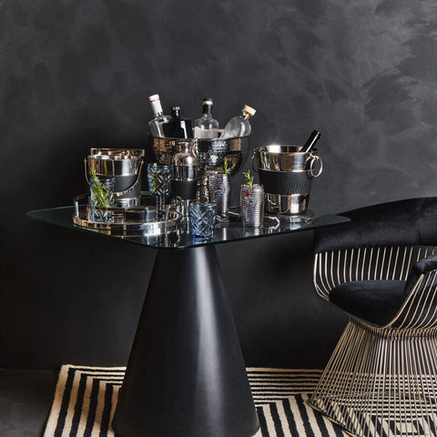 Black glass-top table with sleek tapered pedestal base next to mixology silver ice bucket with silver finish and tongs