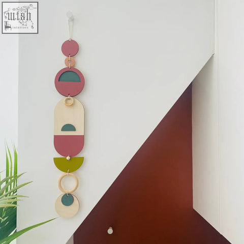 A modern wall art piece featuring layered wooden geometric shapes in muted pink, green, beige, and teal tones