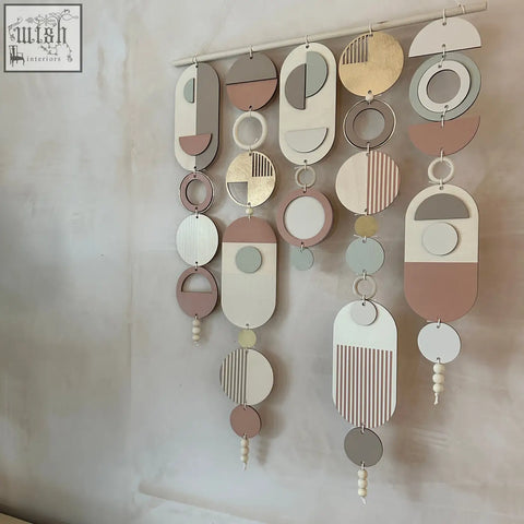 A modern wall art piece with a gold metal hoop and layered wooden shapes in blush pink accents and muted tones