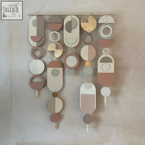 A modern wall art featuring layered wooden discs in beige gray with pink accents and gold metal hoop detailing