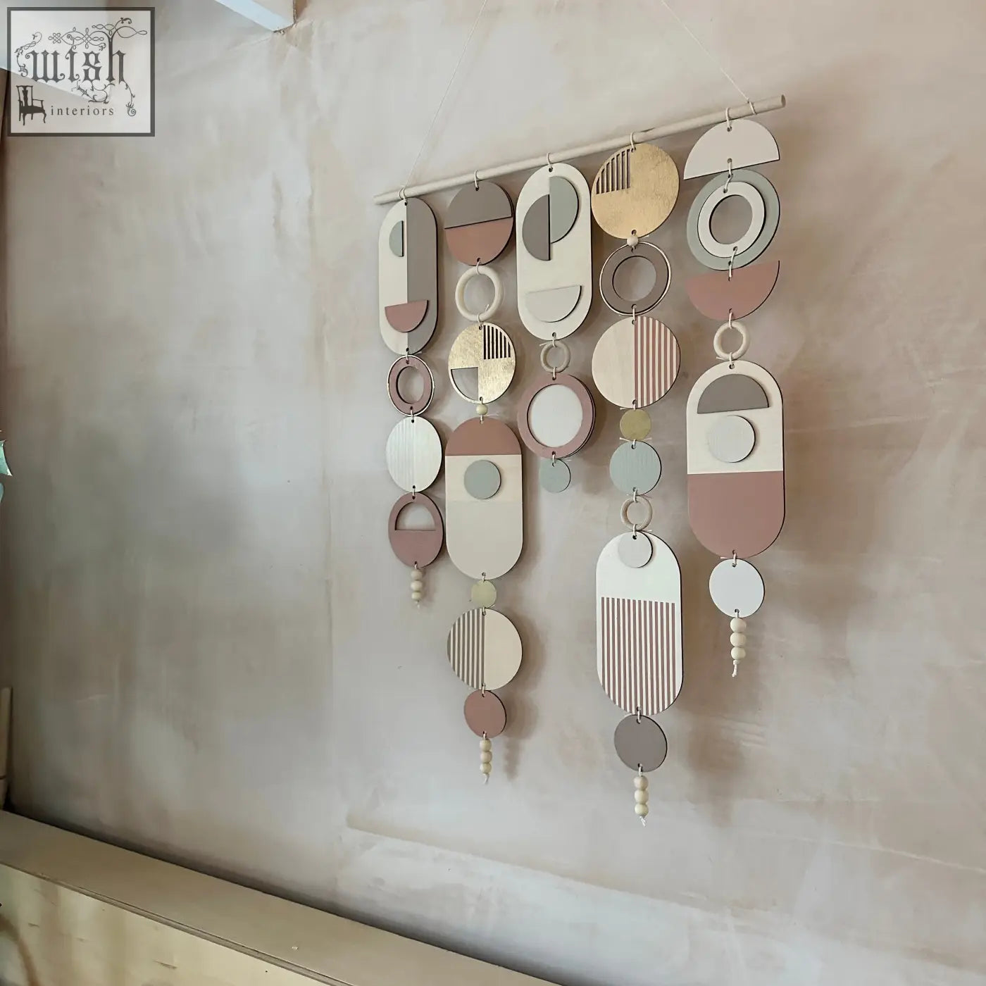 Modern wall art featuring layered wooden shapes with pink accents and gold metal hoop detailing in soft neutral tones