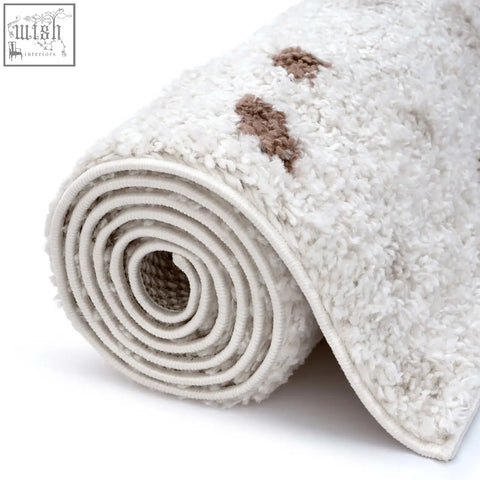A plush moodi shaggy cream bath mat with soft texture and two small brown embroidered patches in cream terra biscotti