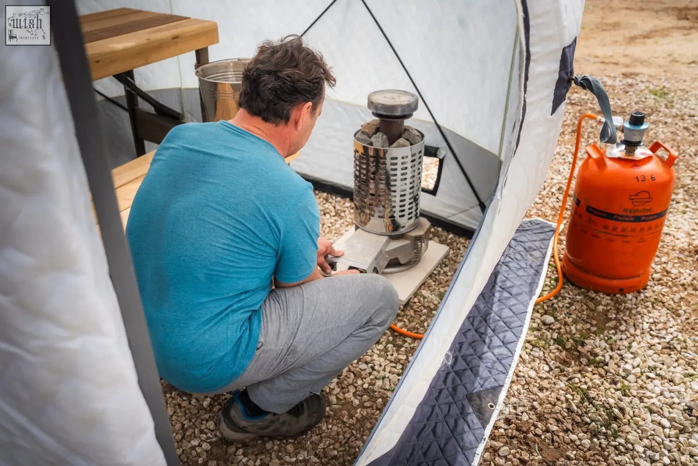 Silver cylindrical camping stove with mesh body and black handle connected to an orange propane tank for r14s safety pack second tent