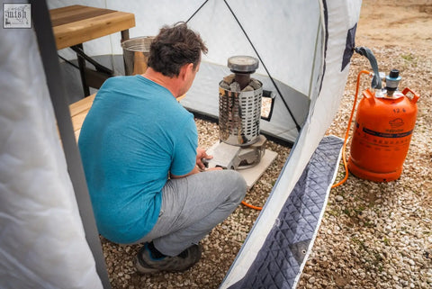 Silver cylindrical camping stove with mesh body and black handle connected to an orange propane tank for r14s safety pack second tent