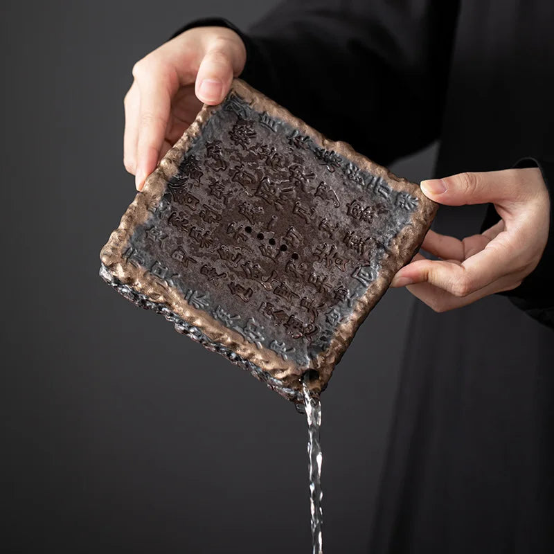 A rustic dark bronze ceramic tile with intricate engraved patterns and rough textured edges from ocha set