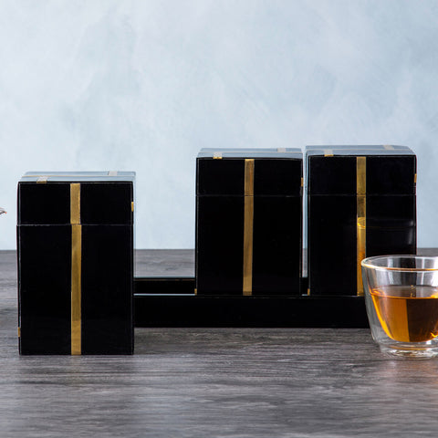 Three sleek odell black glossy black resin tea boxes with gold set stripes on dark wood beside glass cup of amber tea