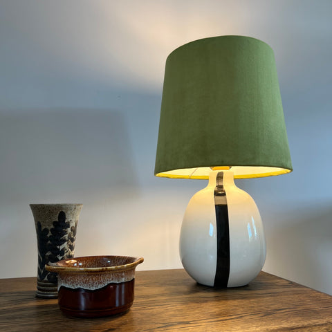 A sleek white ceramic table lamp with a bold black stripe and an olive green velvet french drum lampshade