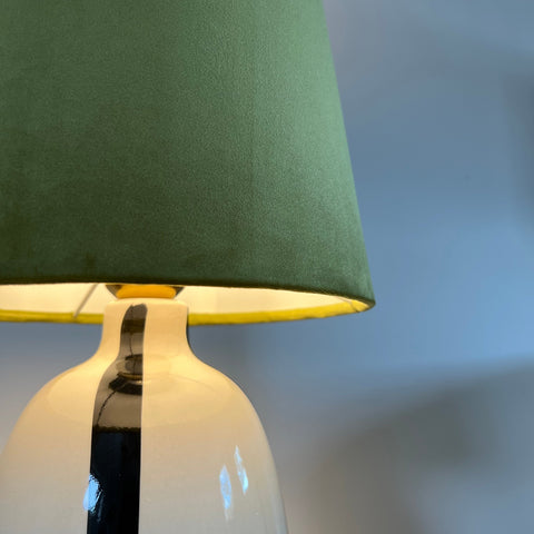 A sleek white ceramic table lamp with a bold black stripe and an olive green velvet french drum lampshade