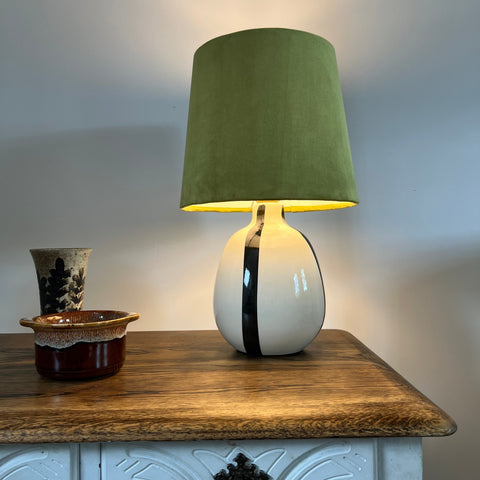 A sleek white ceramic table lamp with a bold black stripe and an olive green velvet french drum lampshade