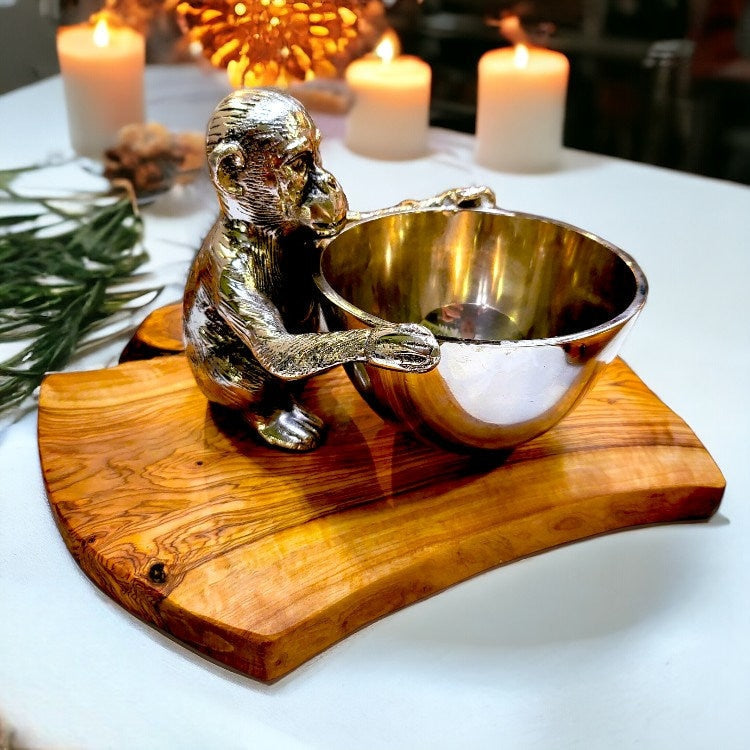 Silver monkey figurine holding a polished brass bowl on an olive wood base serving board with olive tree grain