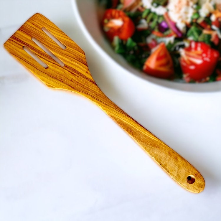 A wooden slotted spatula with a natural golden-brown olive wood finish showing olive trees story grain
