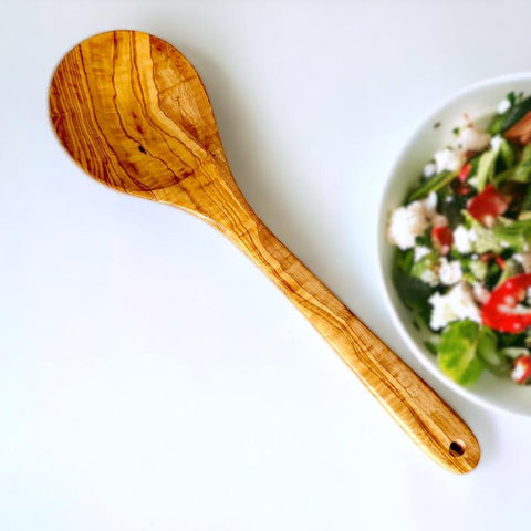 A rustic olive wood cooking spoon with warm golden-brown finish and wavy grain from olive trees story