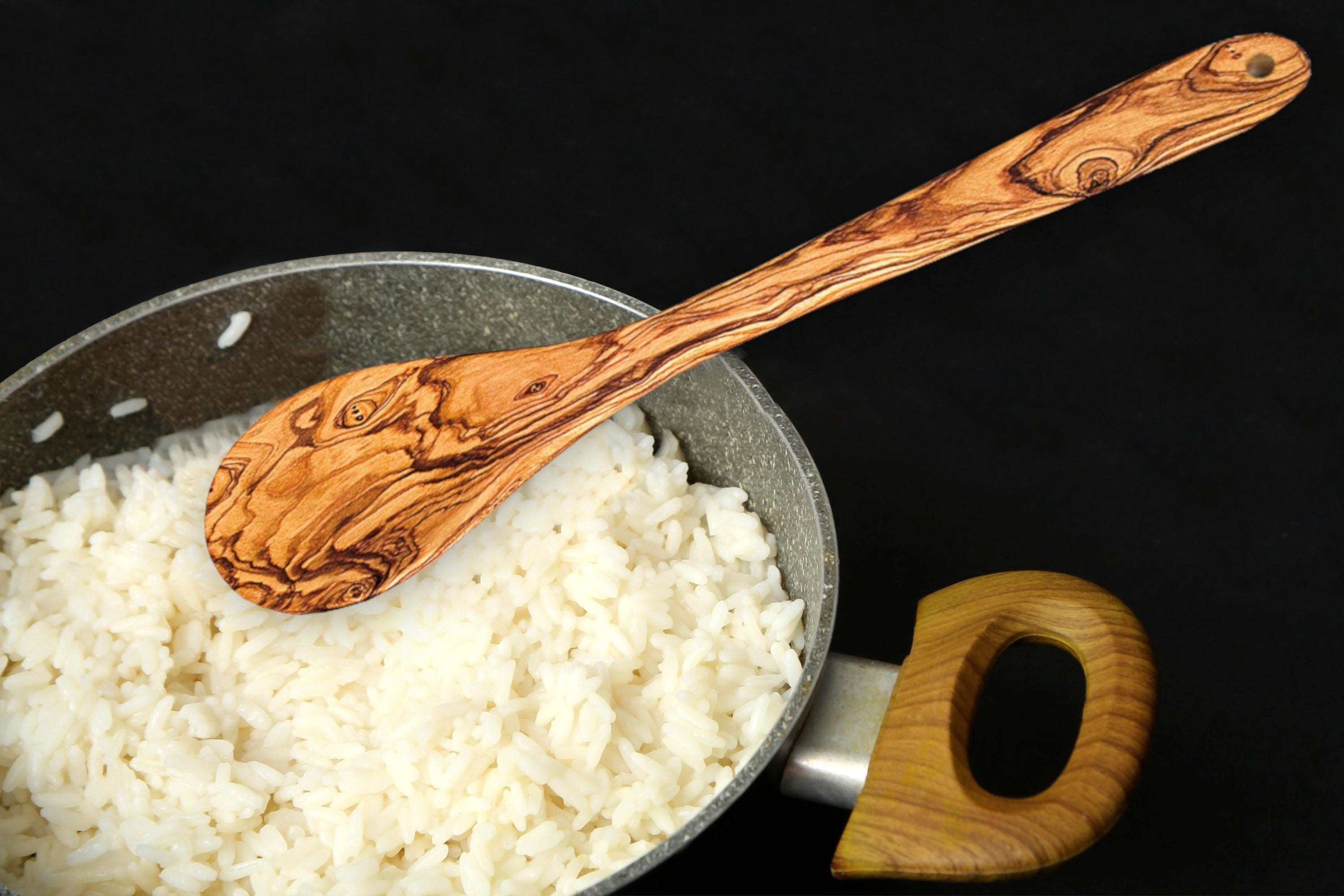 A warm brown olive wood cooking spoon with natural swirling grain rests in a pan of white rice for serving