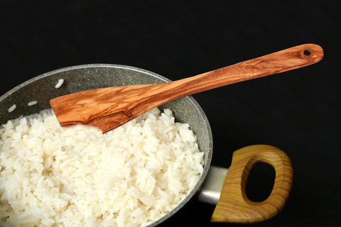 A natural olive wood flat curved spatula 30 cm with warm brown finish resting in a pan of cooked white rice