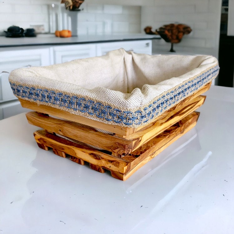 A rustic olive wood bread basket with a beige linen-lined interior and blue embroidered trim 28 cm rectangular shape