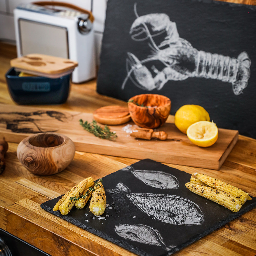 Black slate serving board with white fish etching holding grilled corn beside olive wood salt pot for kitchen use