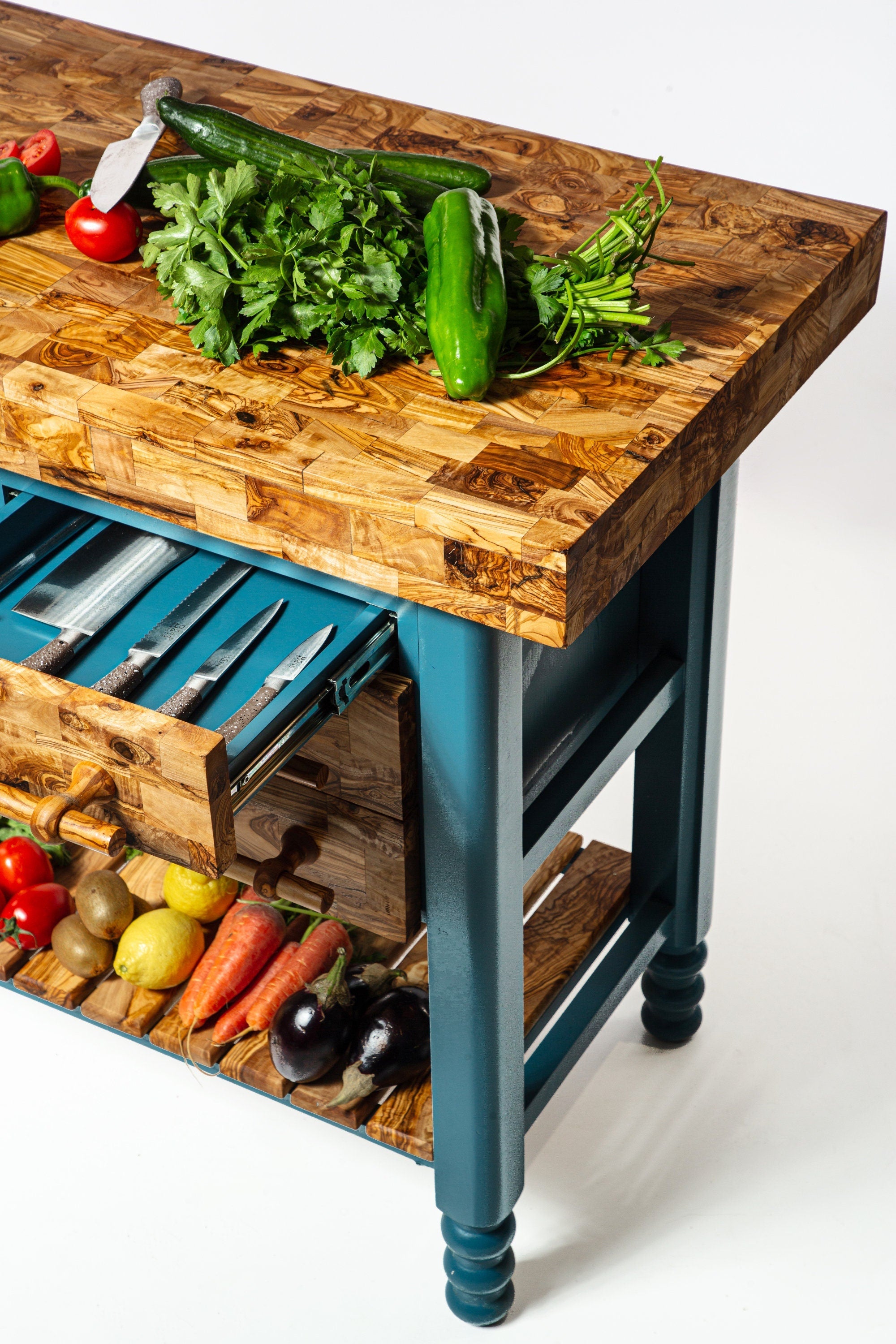 Rustic olive wood table butcher block kitchen island with teal legs and drawers holding knives topped with fresh veggies