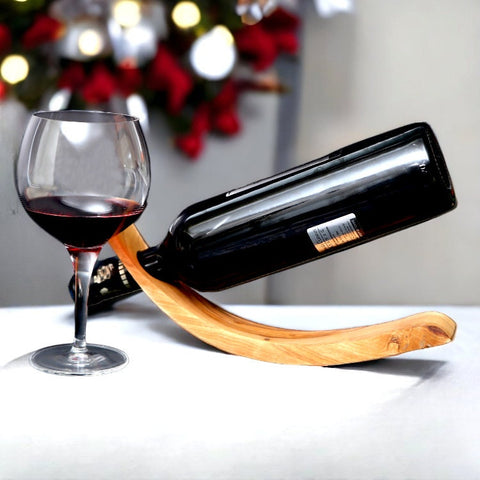 A sleek black wine bottle rests diagonally on a curved olive wood wave bottle holder next to a filled glass of red wine