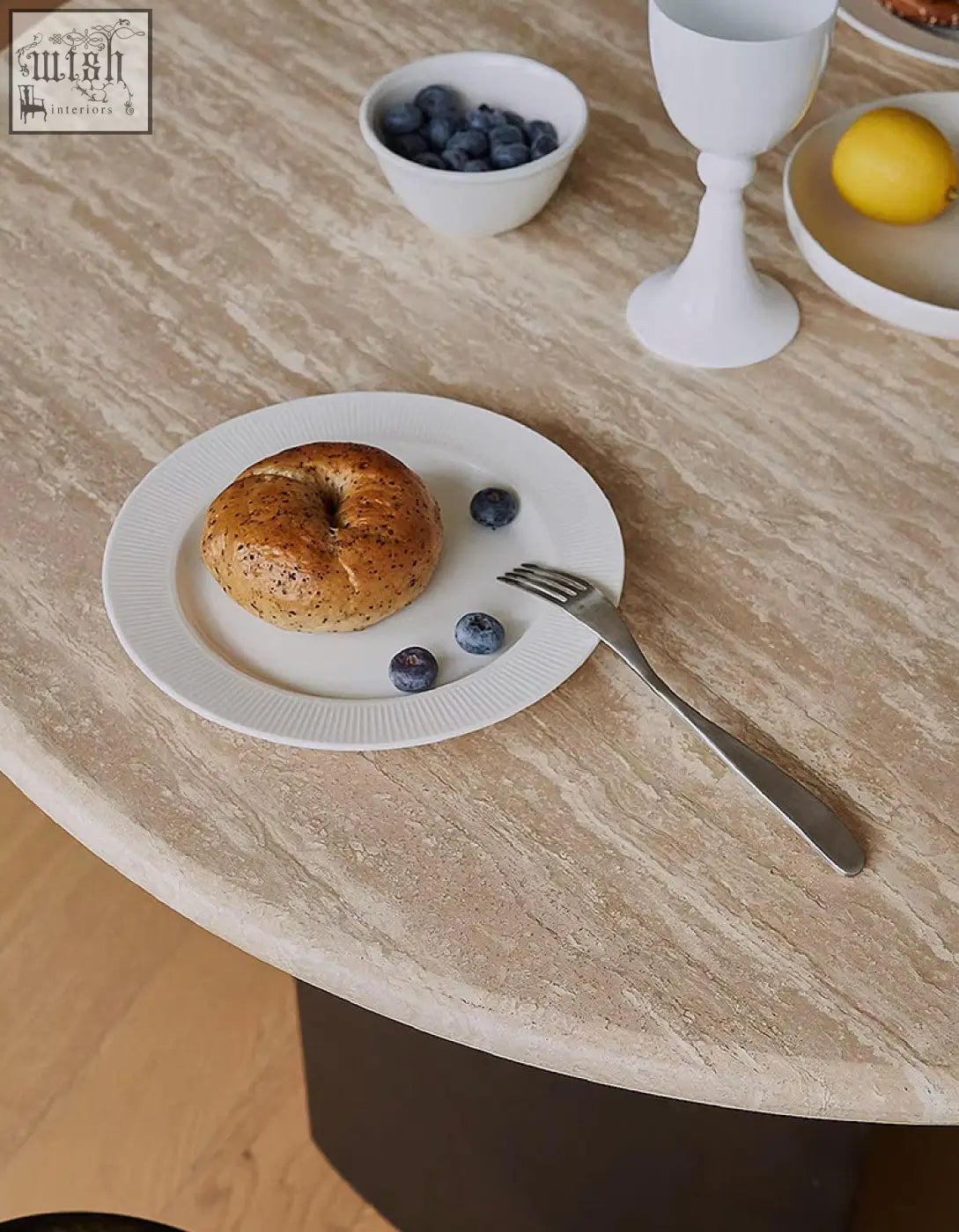 A golden-brown bagel with poppy seeds on a white plate with blueberries and a fork on an oval dining table