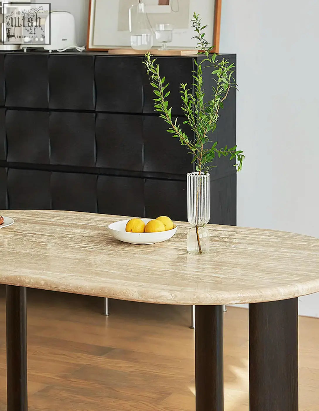 A tall glass vase with greenery sits on the travertin and solid wood o malley oval dining table