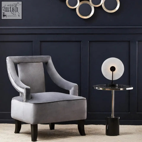 Gray velvet armchair with curved arms and dark wooden legs next to a black marble end table with stainless steel base