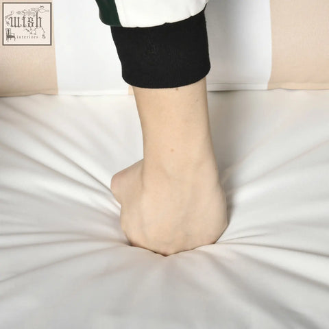 A foot in black ribbed sock rests on white fabric near outsunny light grey rattan coffee table with stylish striped bolster cushion