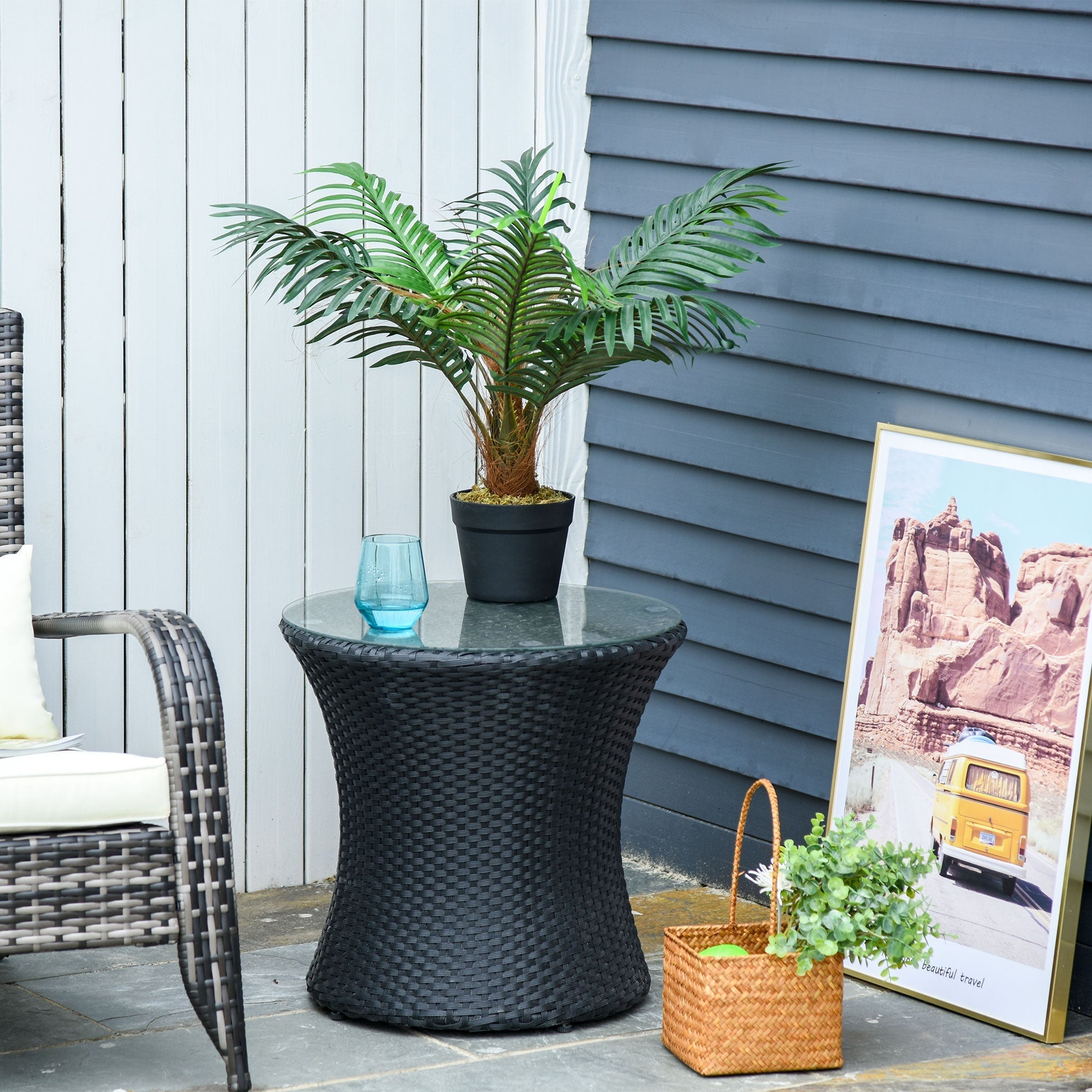 Black woven rattan side table with glass top next to an artificial palm plant in a cement-filled pot for indoor decor